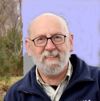Jim is dressed in a blue sweater and a beige shirt. He is wearing his glasses. There is an outdoor scene behind him of mainly blue sky