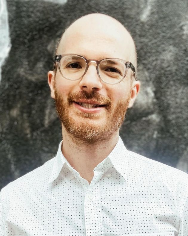 A man smiles directly into the camera. He is bald. His reddish-brown mustache and beard, flecked with hints of white, is worn close to his face. He wears a white button-down shirt featuring a polka dot pattern. His eyeglasses are dark gray acetate and feature rounded rectangular lenses. He is positioned before a gray and white marble backdrop.