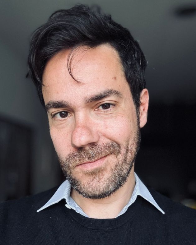 A man smiles wryly into the camera. His black wavy hair is short and brushed away from his forehead. His mustache and beard, also black, are closely cropped to his face and speckled with hints of gray. He wears a blue oxford shirt beneath a navy blue crewneck sweater.