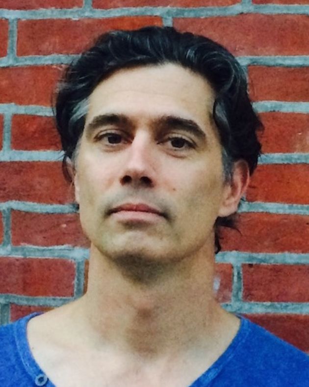 A man stands before a red brick wall. He looks directly into the camera. His wavy black hair is short and brushed back from his face. It is punctuated by gray at the temples. He wears a bright blue scoop-neck tshirt.