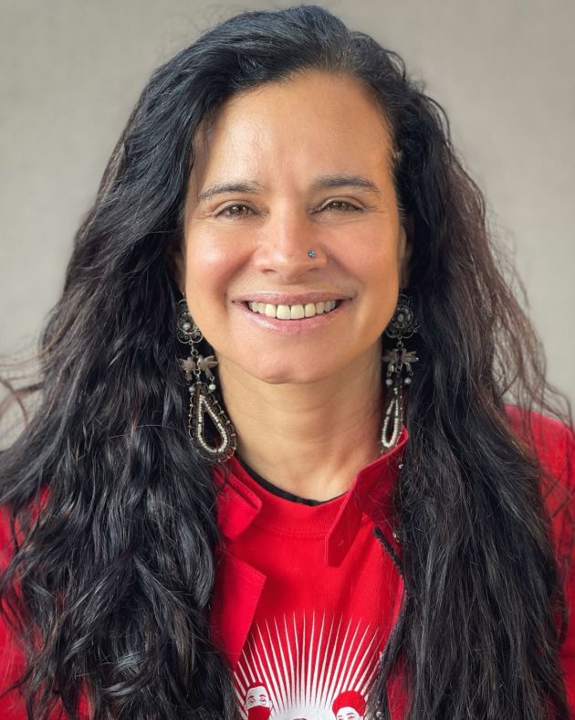 A woman smiles broadly into the camera, her wavy dark brown hair cascading over her shoulders. She wears large teardrop-shaped earrings with small white and black beads. Her nose is pierced with a small turquoise stud. She wears a bright red tshirt with a white graphic and a bright red jacket. She is positioned before a beige background.
