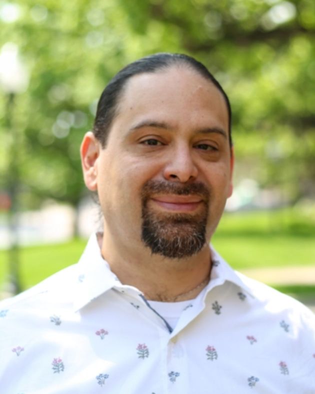 A man stands outside surrounded by lush green trees. He looks directly into the camera with a soft smile. His black hair is slicked back from his face. His mustache and goatee, also black, are worn short and peppered with hints of gray. He wears a white tshirt beneath a white oxford shirt that features flower sprigs of various colors.