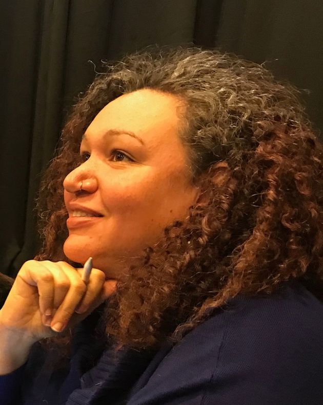 A woman is smiling into the distance, her head resting on her right fist while it grips a pencil. She is in profile. Her shoulder-length hair is curly and brown, with hints of gray. Her nose is pierced with a small silver hoop. She wears a navy jacket. Behind her, one can see a black backdrop.