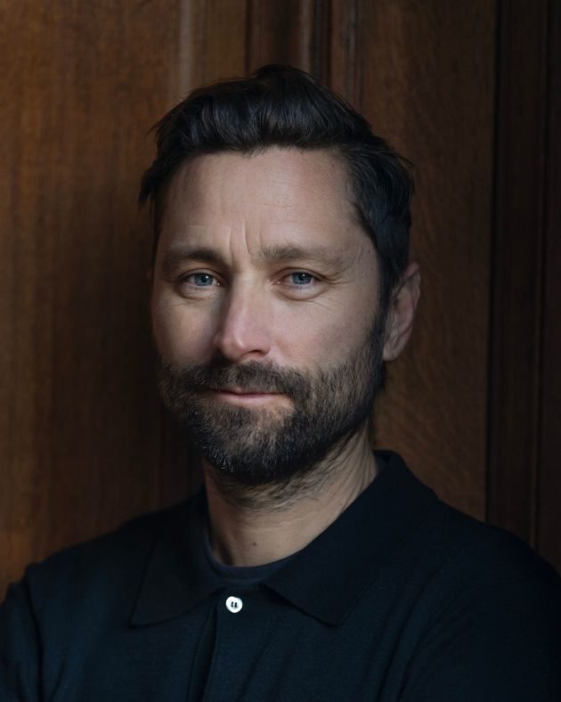 A man smiles gently at the camera. He stands before warm wood panelling. He has short, dark brown hair that his swept back from his forehead. His mustache and full beard are also dark brown. He wears a navy blue tshirt beneath a navy blue polo shirt.