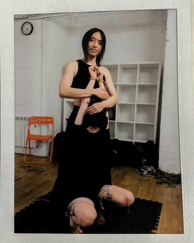 June is holding Sara's arms above their head, with Sara forced into a kneeling position by ties around both legs.