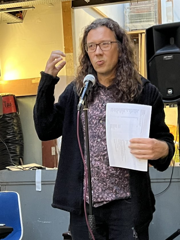 A man with long hair, glasses, and a black cardigan gesticulates in front of a microphone