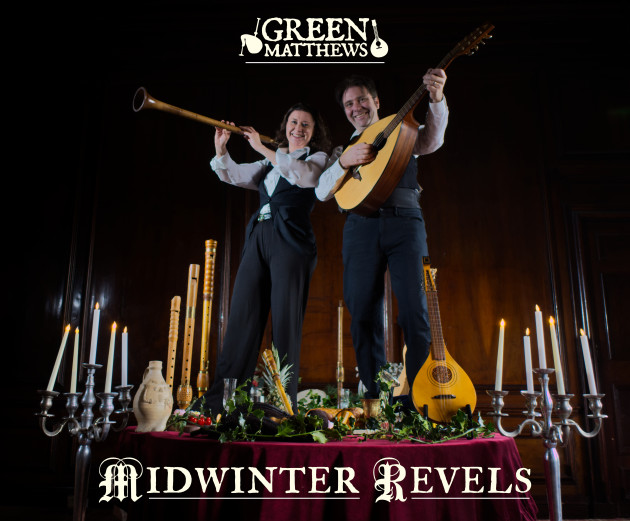 Photo of GreenMatthews - two performers, one male and one female - holding musical instruments whilst standing on a tablet laden with food, like a banquet.