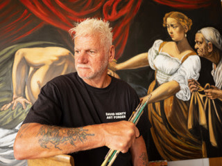 David Henty holding paint brushes and standing in frront of a painting David Henty holding paint brushes and standing in frront of a painting