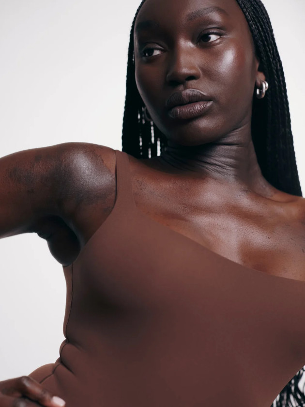 Black female model posing with hand on waist and wearing a brown bodice. Glowy natural skin make up finish and shiny lip gloss.