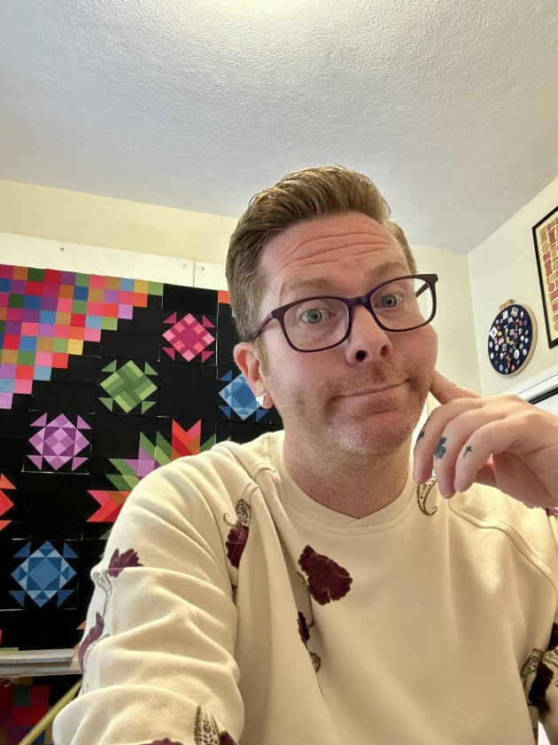 James sitting in front of one of his quilts wearing a cream colored sweater and glasses