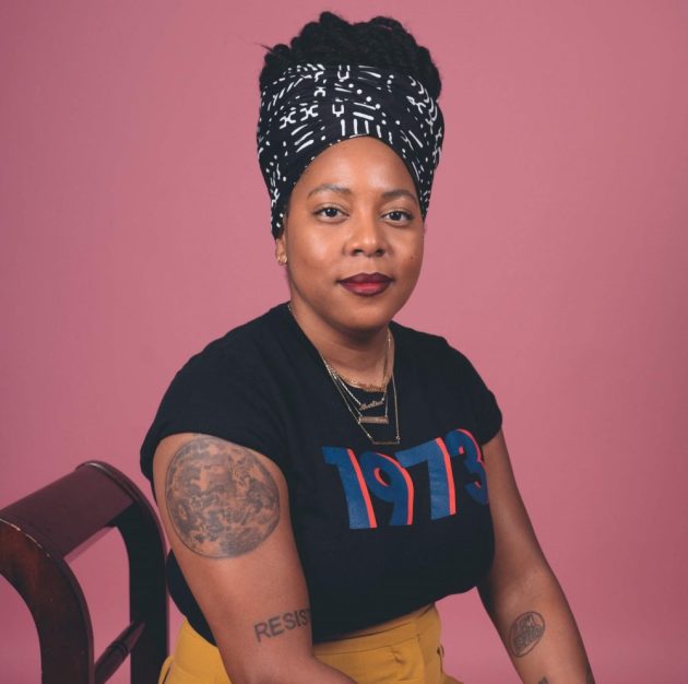Kwajelyn Jackson is sitting on a chair, looking at the camera wearing a black shirt with the words 1973. Her hair is wrapped. She has tattos in her right shoulder and forearms as well as her right forearm.